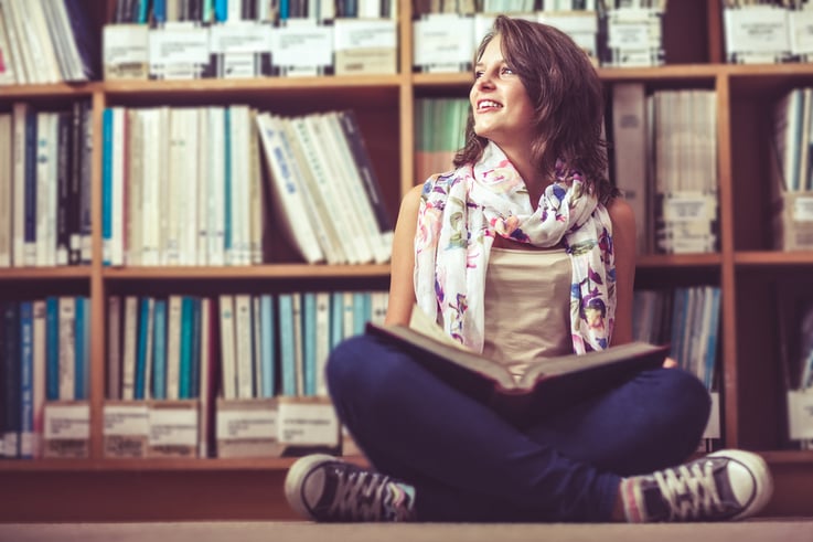 Thoughtful female student sitting against bookshelf with a book on the library floor